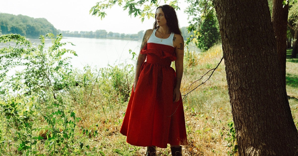 Image of Waxahatchee in a red dress on the bank of a river surrounded by greenery